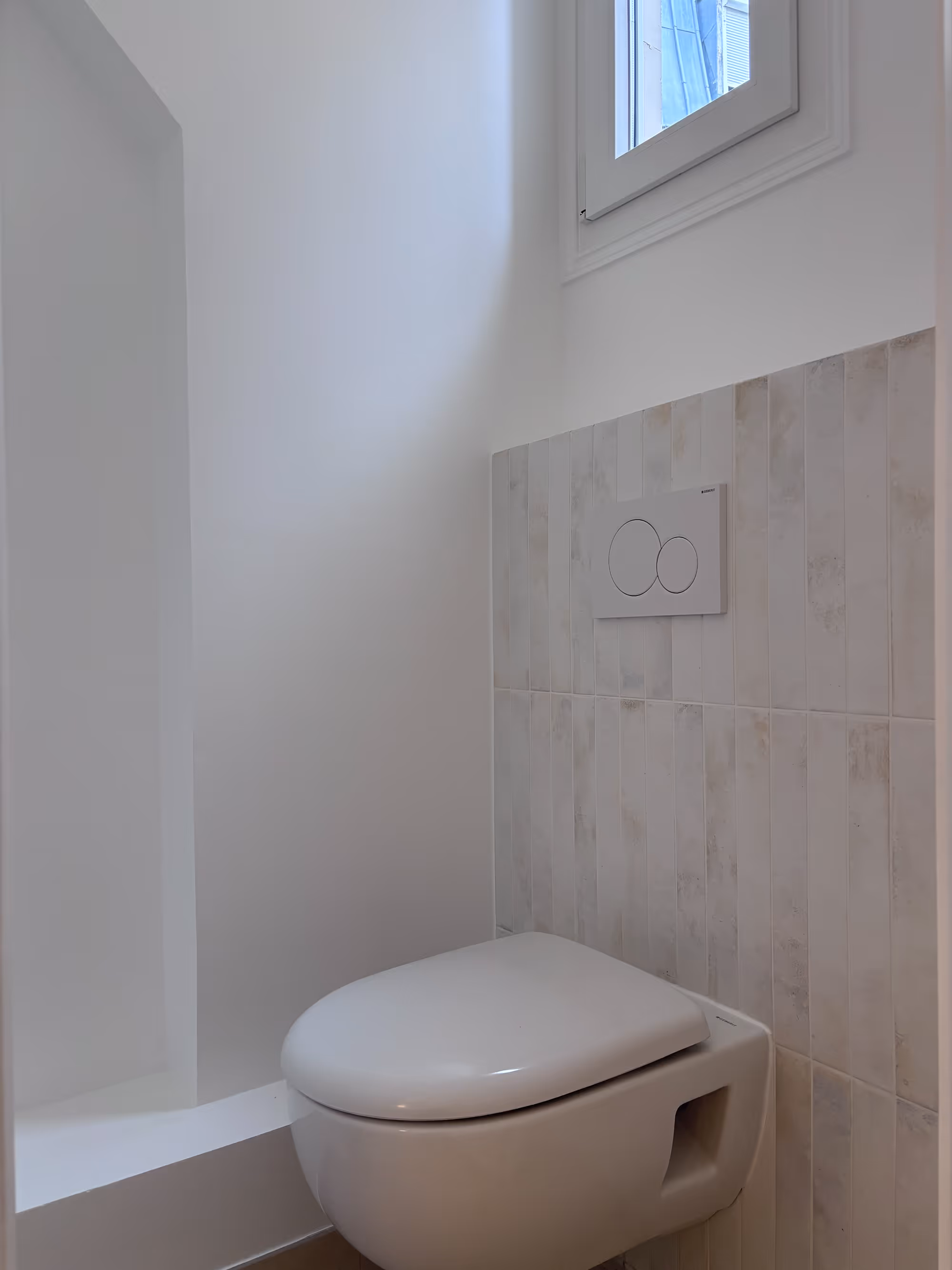 Rénovation d'un appartement haussmannien parisien (5e arr.) : toilettes avec WC suspendu, carrelage mural beige en format vertical, plaque de chasse double touche et petite fenêtre en hauteur. Réalisation : Aude Riou, architecte d'intérieur.