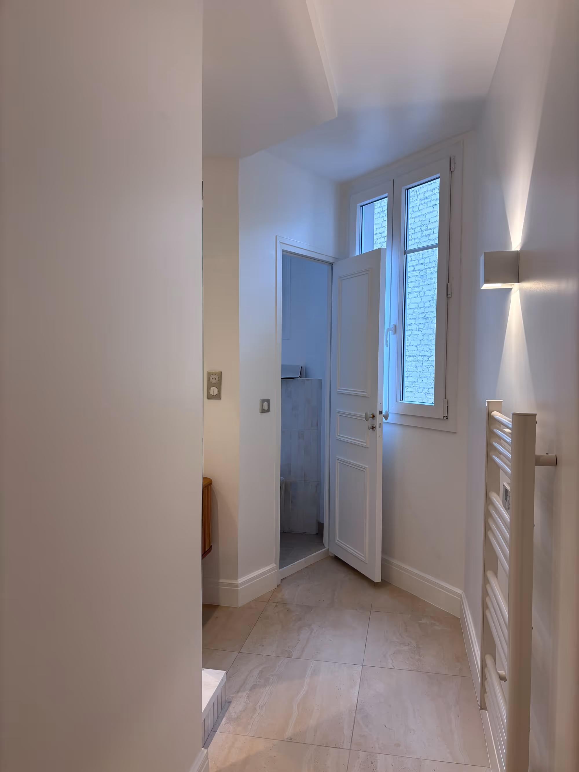 Rénovation d'un appartement haussmannien parisien (5e arr.) : salle de bain avec sol en marbre clair, porte à panneaux blanche, fenêtre à petit bois, applique murale rectangulaire et sèche-serviettes blanc. Réalisation : Aude Riou, architecte d'intérieur.