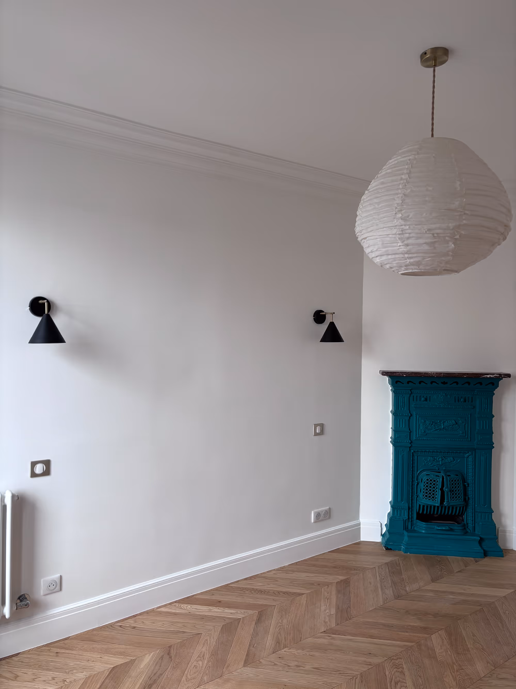 Rénovation d'un appartement haussmannien parisien (5e arr.) : chambre avec murs blancs, parquet en chevrons, corniche de plafond, deux appliques coniques noires, suspension sphérique en papier et poêle en fonte sculpté bleu canard. Réalisation : Aude Riou, architecte d'intérieur.