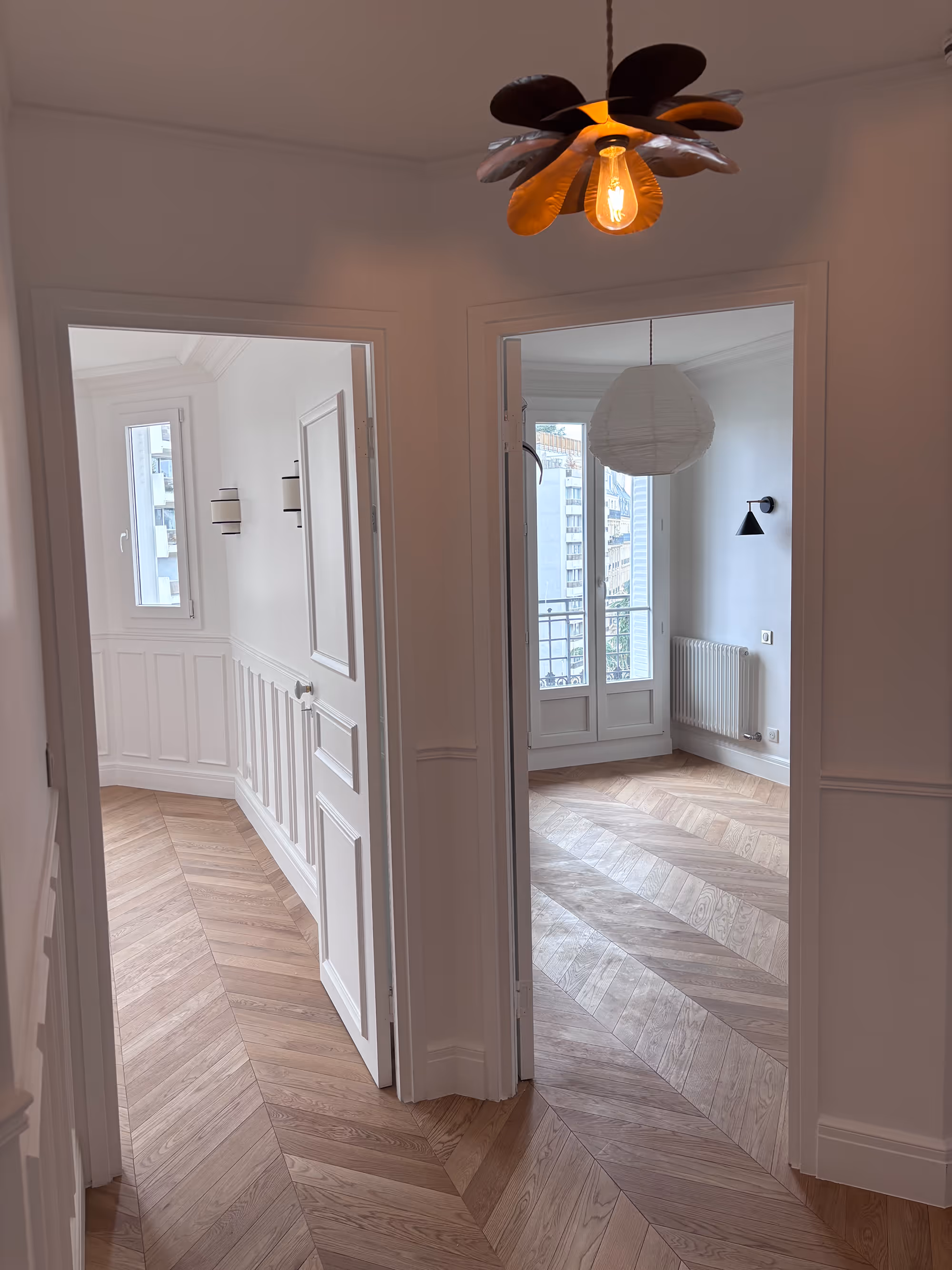 Couloir lumineux d'un appartement haussmannien parisien (5e arr.) : murs blancs, parquet en point de Hongrie, grandes fenêtres et luminaires modernes. Réalisation : Aude Riou, architecte d'intérieur.