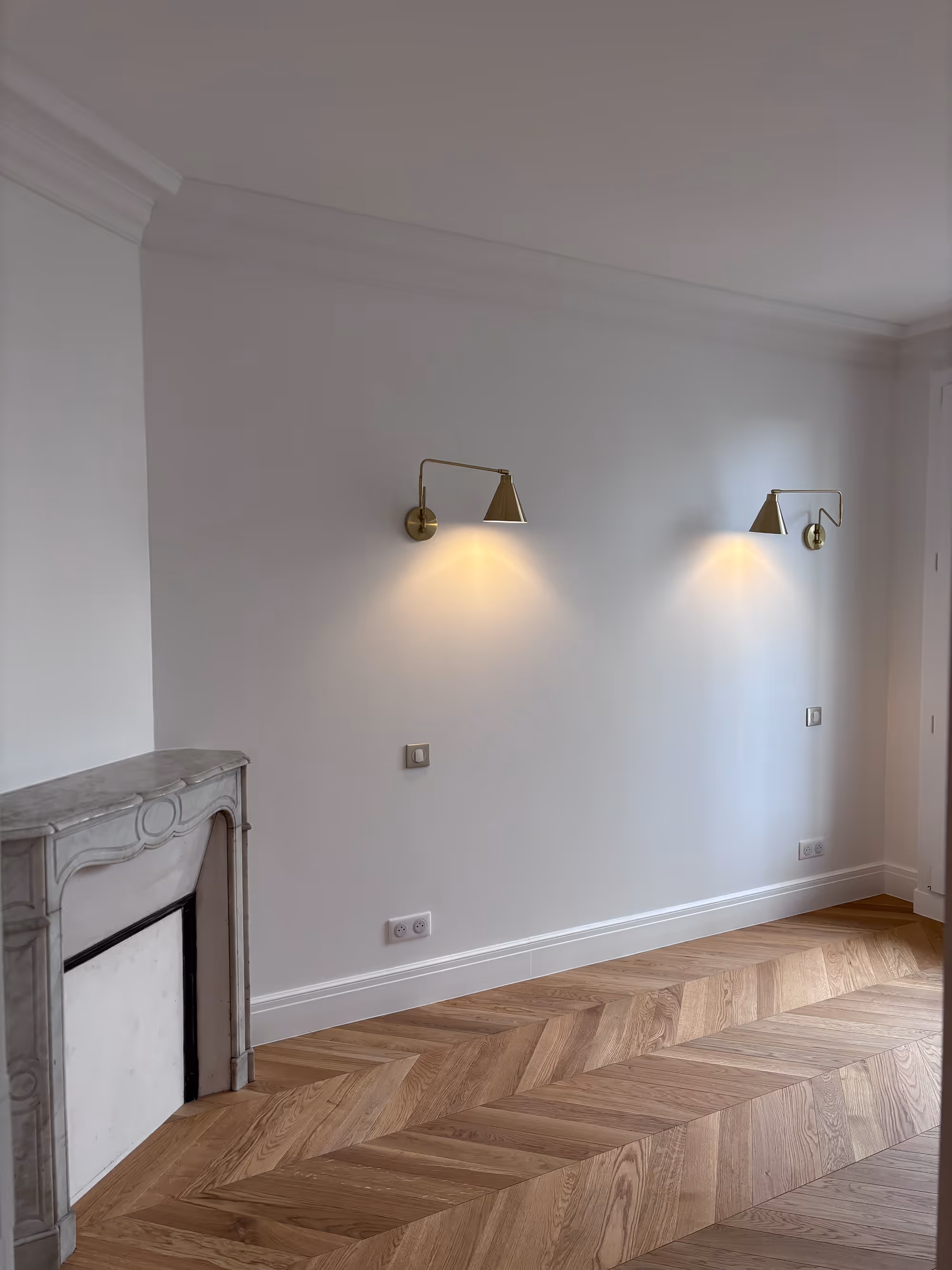 Rénovation d'un appartement haussmannien parisien (5e arr.) : salon avec murs blancs, parquet en chevrons, corniche de plafond, cheminée en marbre sculpté et deux appliques orientables en laiton doré. Réalisation : Aude Riou, architecte d'intérieur.