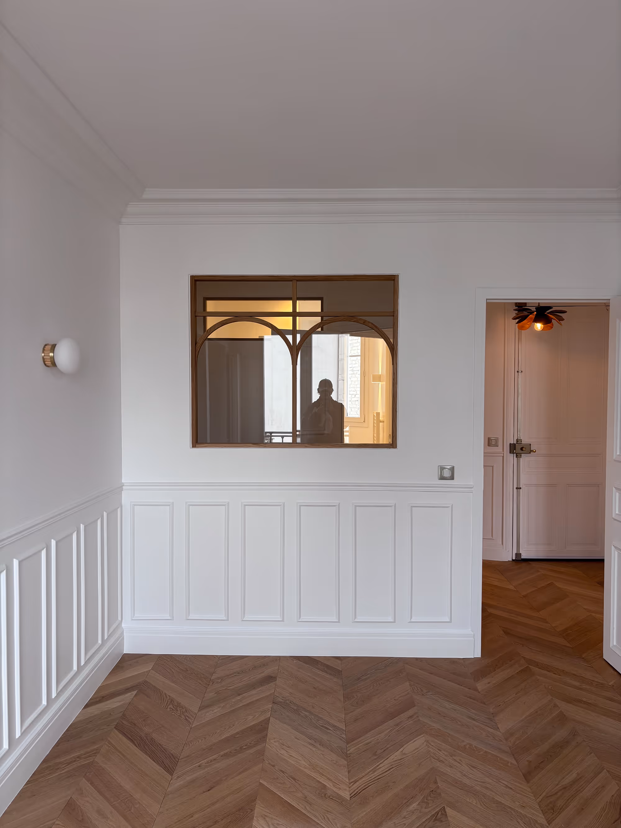 Rénovation d'un appartement haussmannien parisien (5e arr.) : murs à lambris blancs, parquet en chevrons, applique murale et fenêtre à cadres en bois avec arches réalisée sur mesure. Réalisation : Aude Riou, architecte d'intérieur.