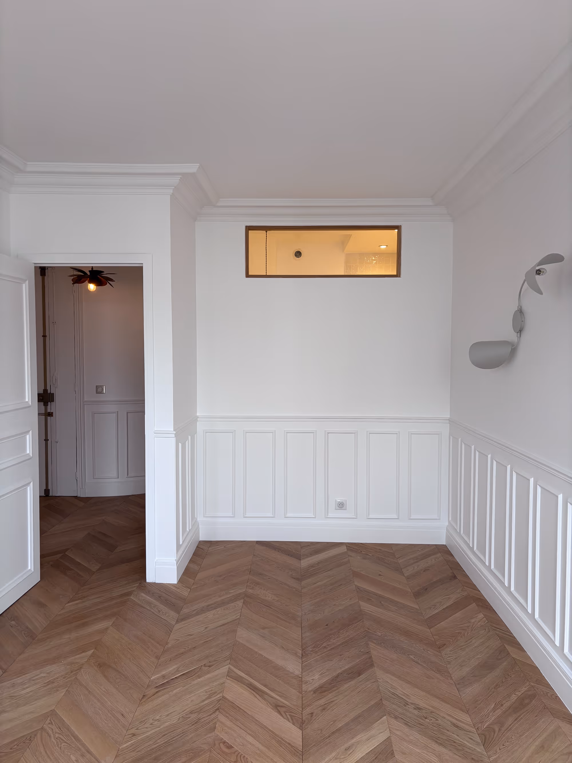 Rénovation d'un appartement haussmannien parisien (5e arr.) : murs à lambris blancs, parquet en chevrons, fenêtre rectangulaire en hauteur, porte ouverte et applique murale moderne. Réalisation : Aude Riou, architecte d'intérieur.