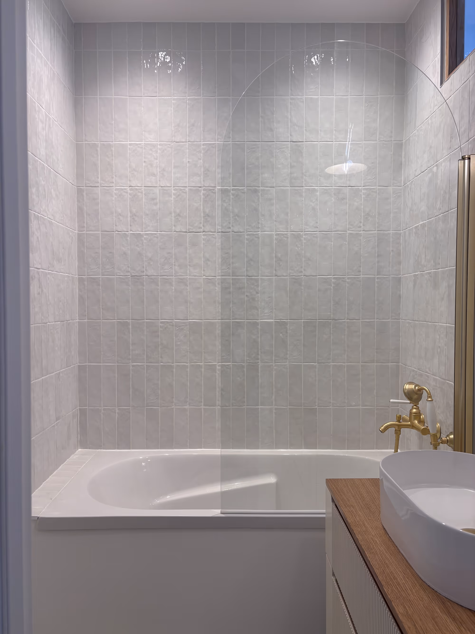 Rénovation d'un appartement haussmannien parisien (5e arr.) : salle de bain avec baignoire blanche, robinetterie dorée, vasque ovale blanche sur plan de travail en bois et carrelage mural gris clair texturé. Réalisation : Aude Riou, architecte d'intérieur.