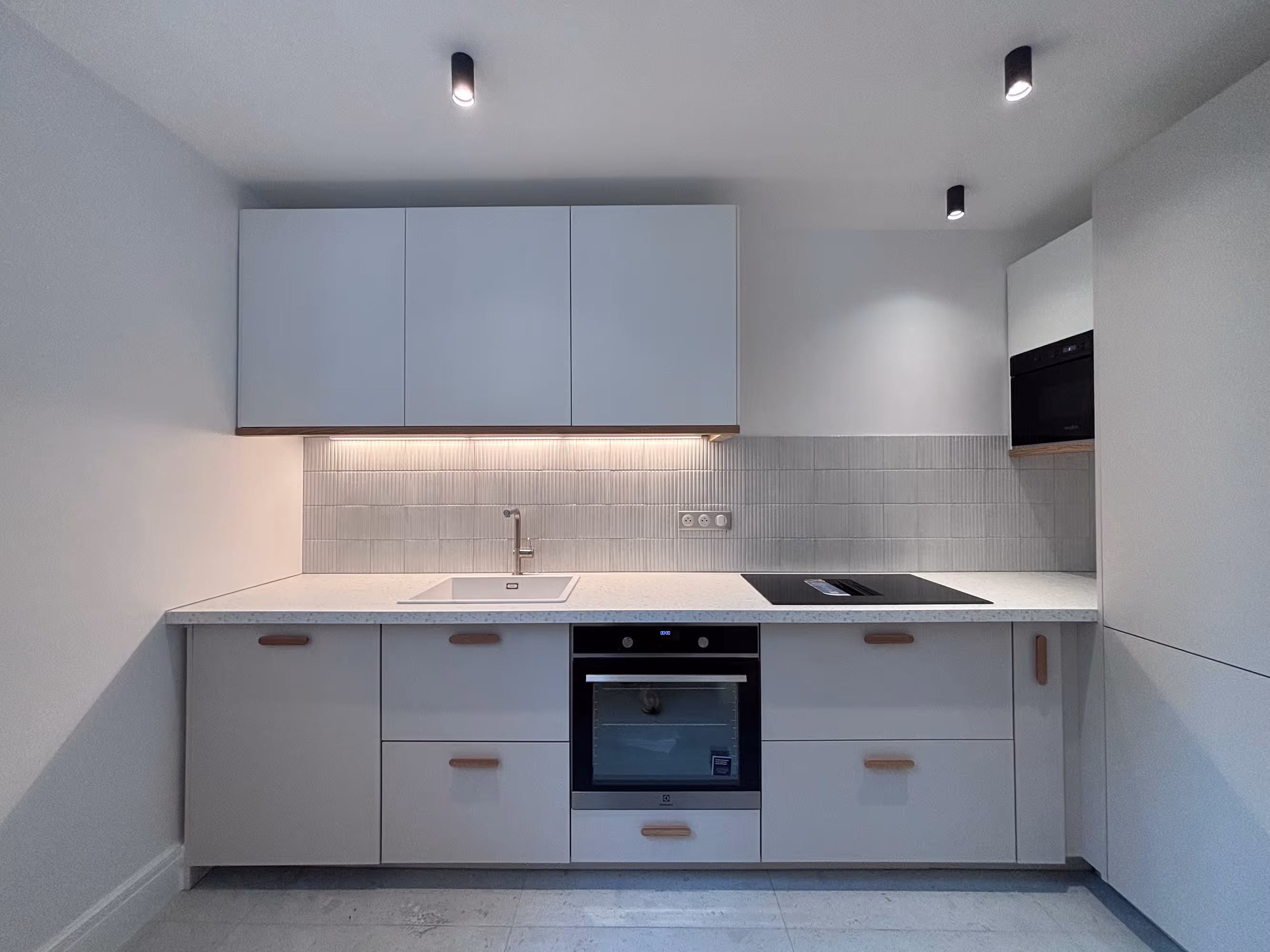 Rénovation d'un appartement haussmannien parisien (5e arr.) : cuisine avec placards gris clair et poignées en bois, plan de travail granité blanc, évier encastré, plaque de cuisson induction, four et micro-ondes encastrés noirs, crédence en carrelage strié vertical et spots noirs au plafond. Réalisation : Aude Riou, architecte d'intérieur.