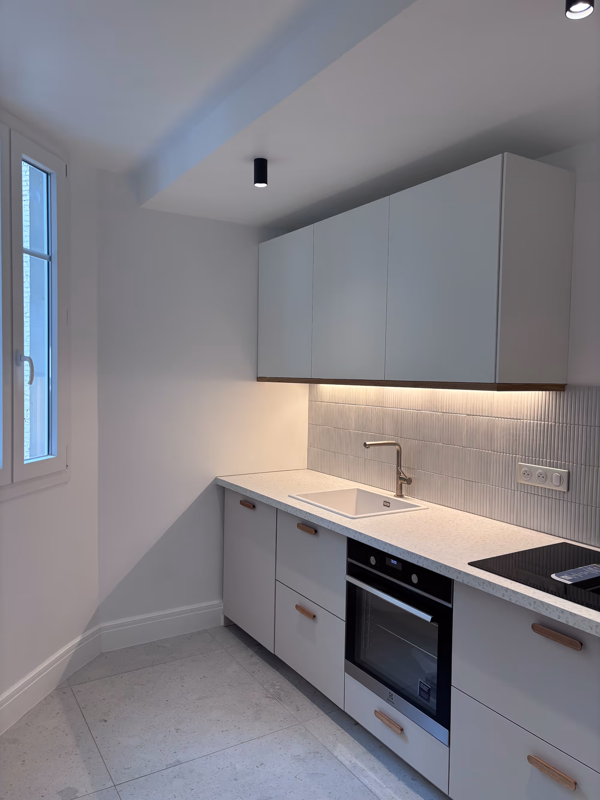 Rénovation d'un appartement haussmannien parisien (5e arr.) : cuisine minimaliste avec placards blancs, four encastré, évier et éclairage sous-meuble. Réalisation : Aude Riou, architecte d'intérieur.