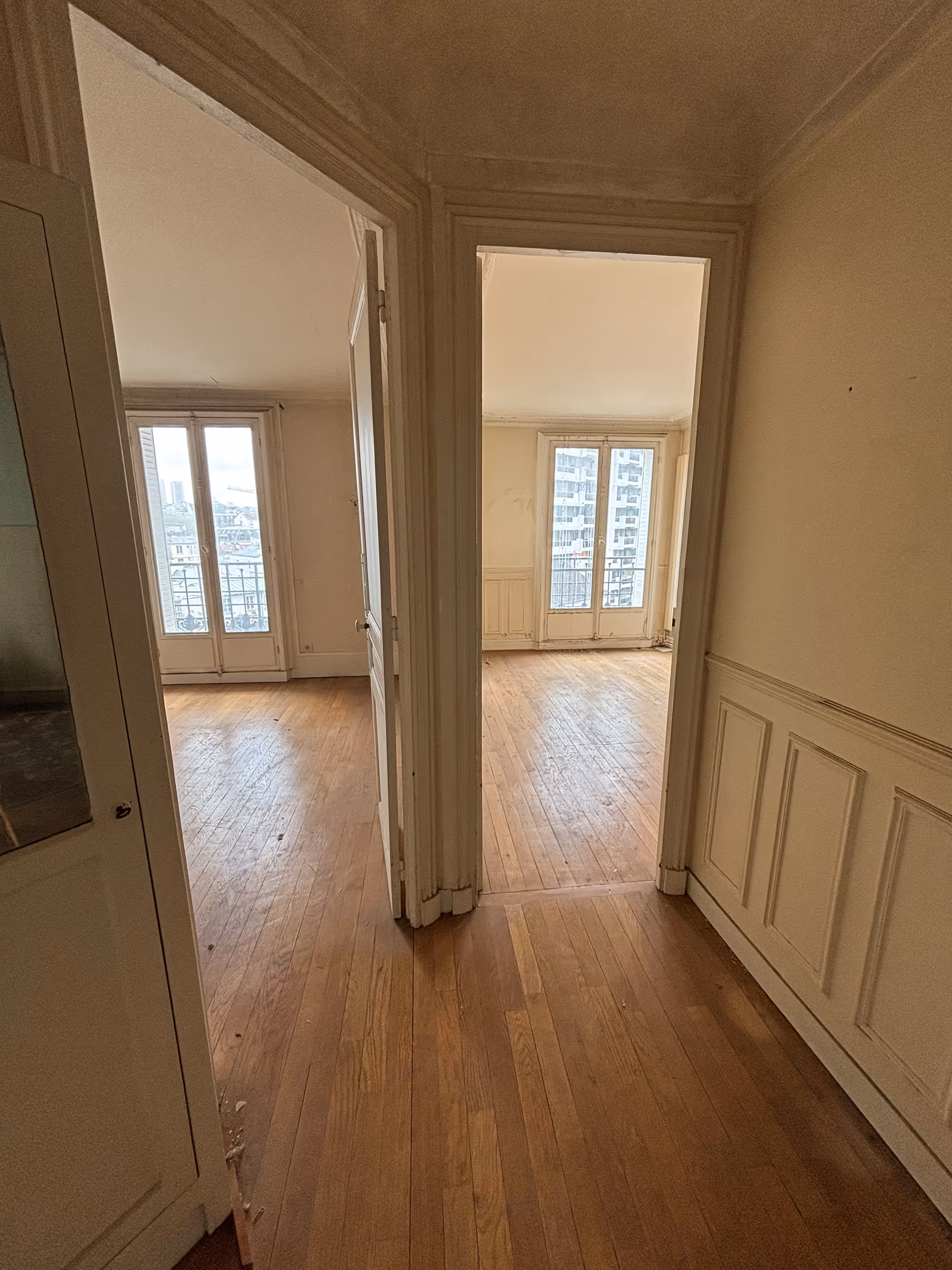 Appartement haussmannien parisien (5e arr.) avant rénovation : couloir donnant sur deux pièces avec parquet usé, lambris à panneaux, corniche de plafond et fenêtres à double vantaux. 