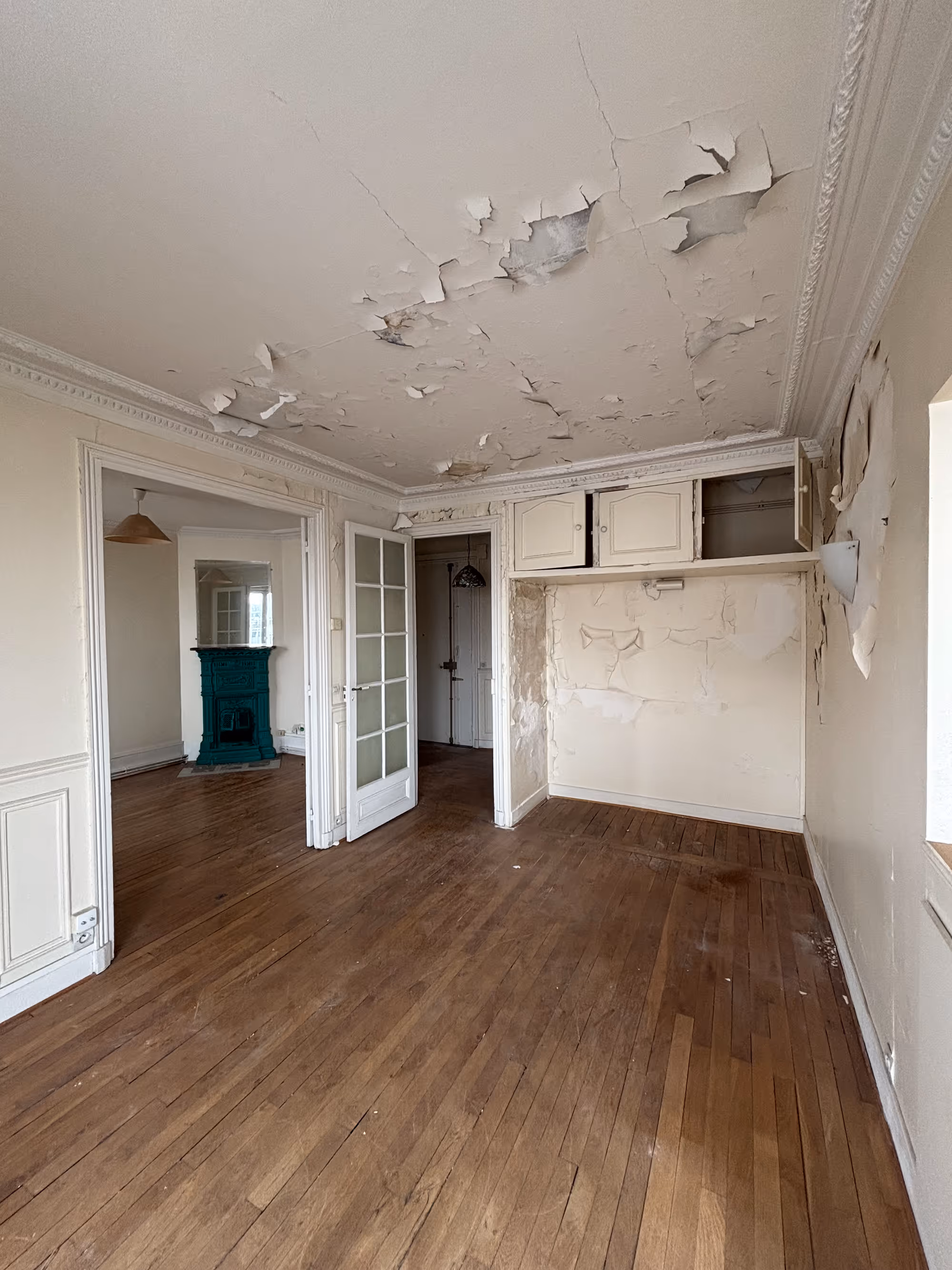 Appartement haussmannien parisien (5e arr.) avant rénovation : pièce de vie avec parquet usé, murs et plafond très dégradés avec peinture décollée, lambris à panneaux, porte vitrée à petits carreaux et poêle en fonte bleu canard en arrière-plan.