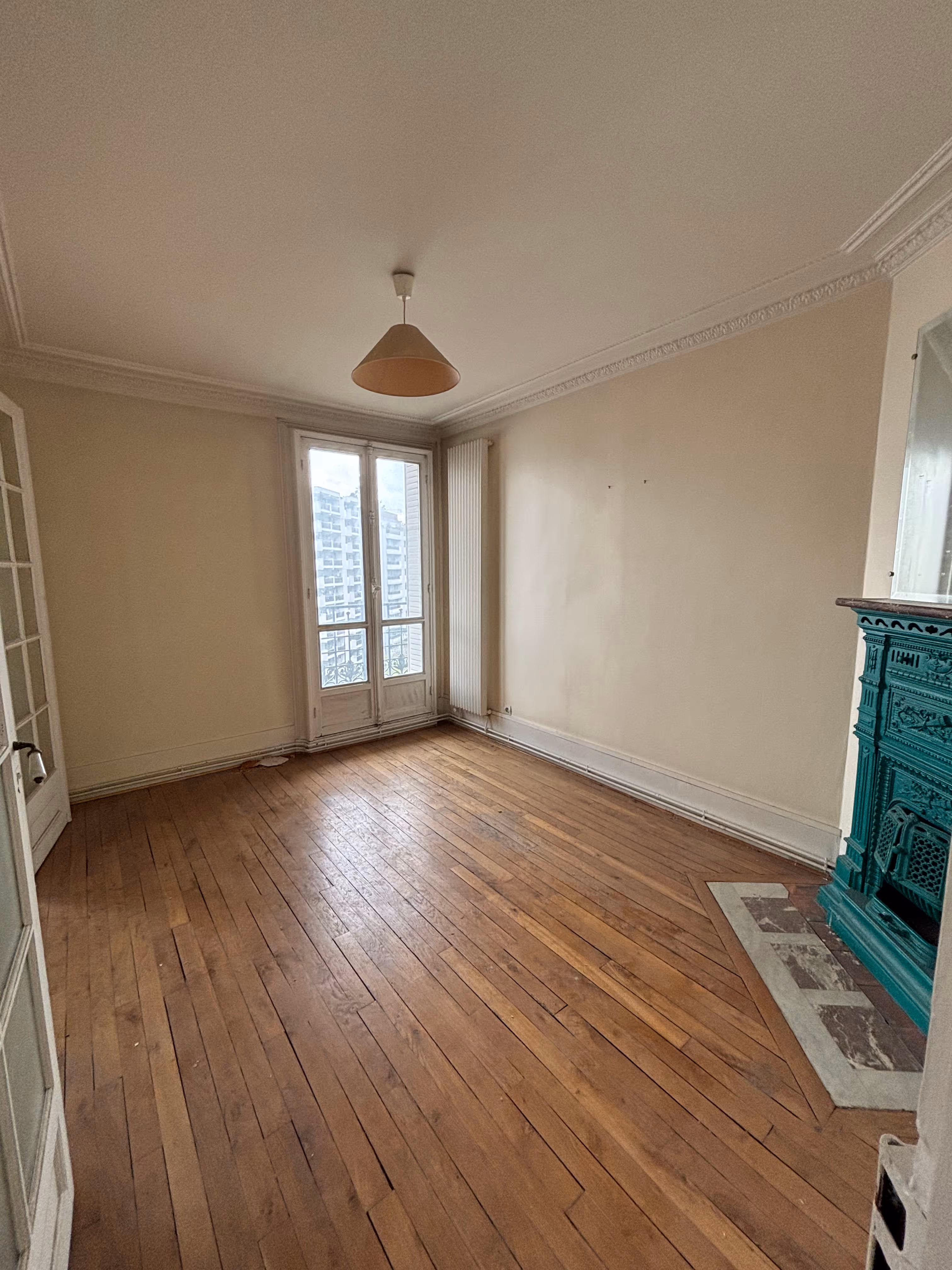 Appartement haussmannien parisien (5e arr.) avant rénovation : chambre avec parquet usé, murs jaunis, corniche de plafond, porte vitrée à petits carreaux, fenêtre à double vantaux et poêle en fonte bleu canard.