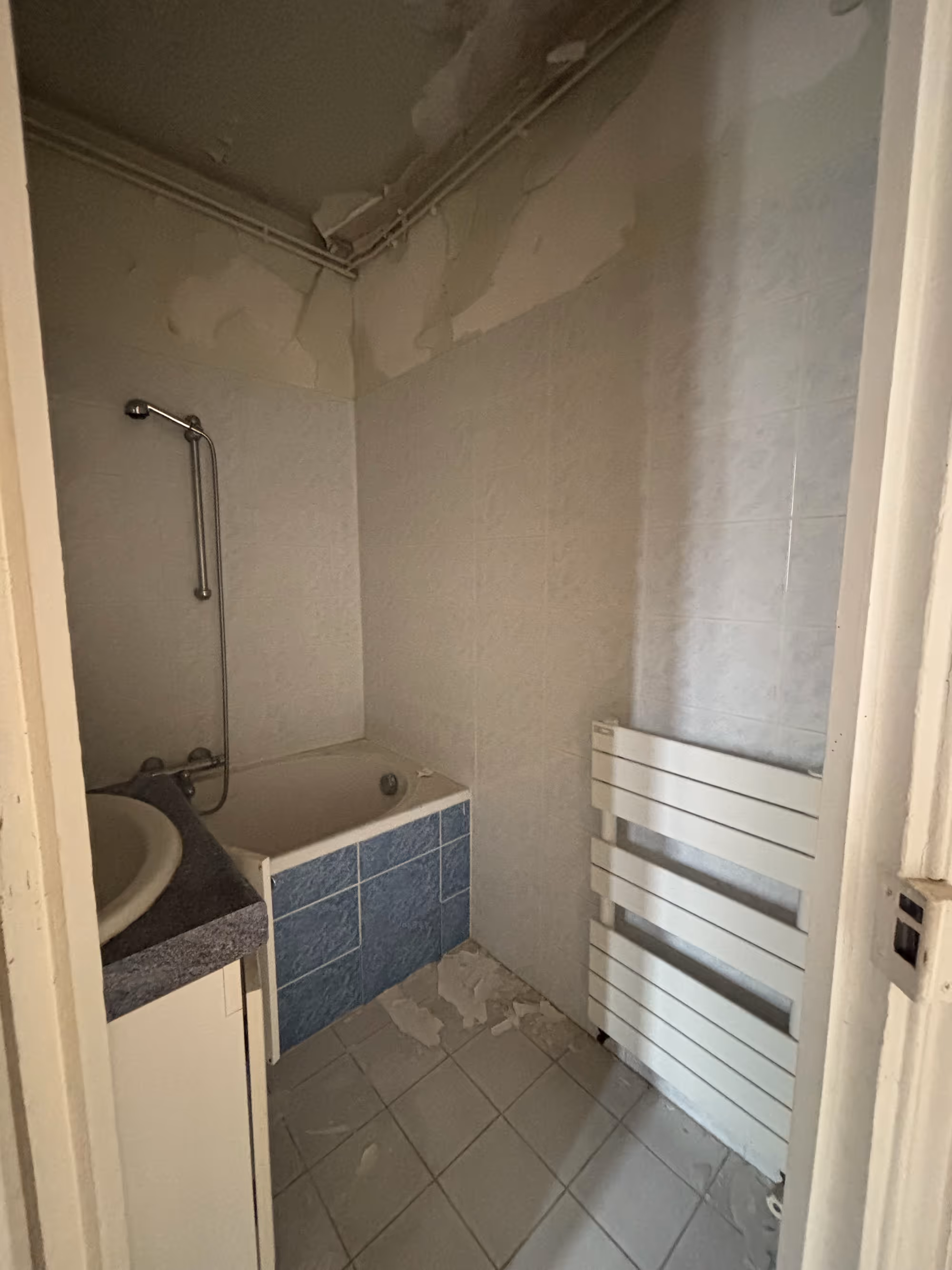Appartement haussmannien parisien (5e arr.) avant rénovation : salle de bain dégradée avec baignoire carrelée en bleu, plafond décollé, sèche-serviettes et carrelage mural usé.
