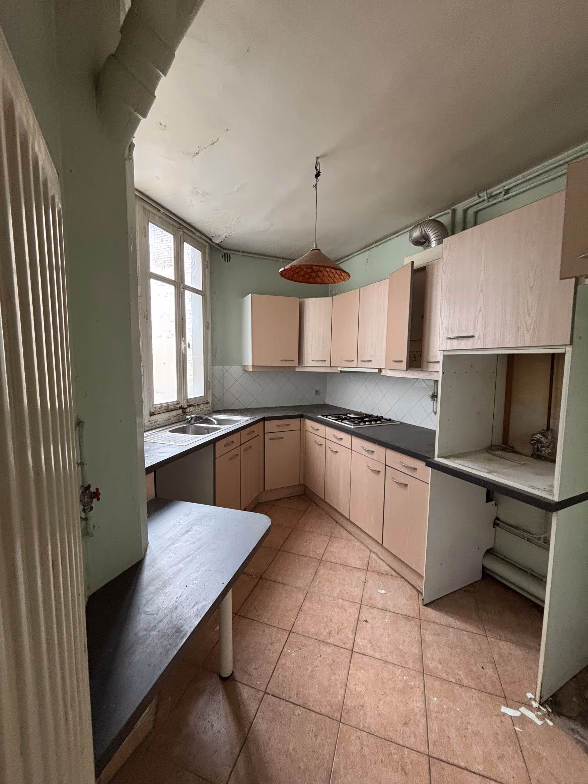 Appartement haussmannien parisien (5e arr.) avant rénovation : cuisine vétuste avec placards roses, plan de travail sombre, carrelage en losanges, sol en carrelage usé et tuyauterie apparente.