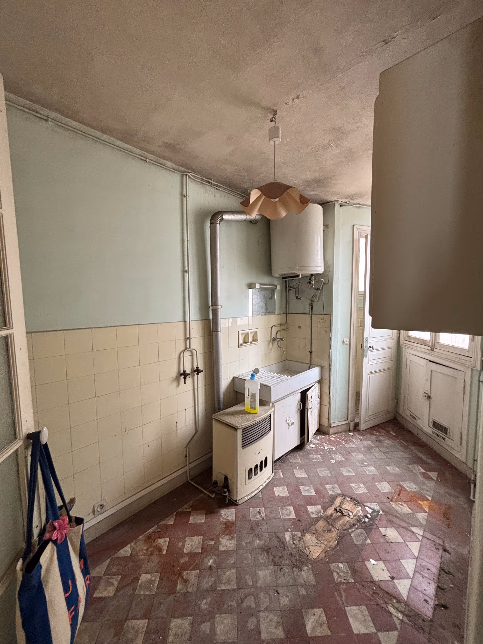 Appartement haussmannien parisien (5e arr.) avant rénovation : arrière-cuisine très dégradée avec carrelage mural ancien, sol en carreaux abîmés, chauffe-eau et tuyauteries apparentes.