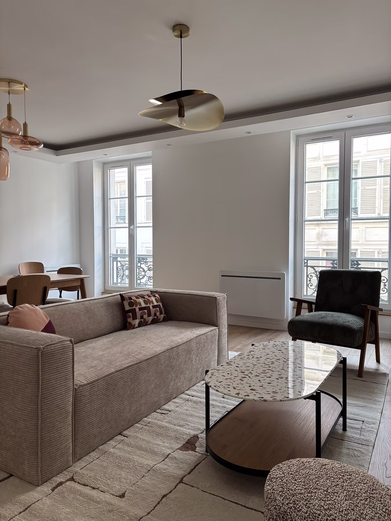 Vue d’ensemble du salon : canapé beige, fauteuil sombre, table basse ronde et grandes fenêtres.