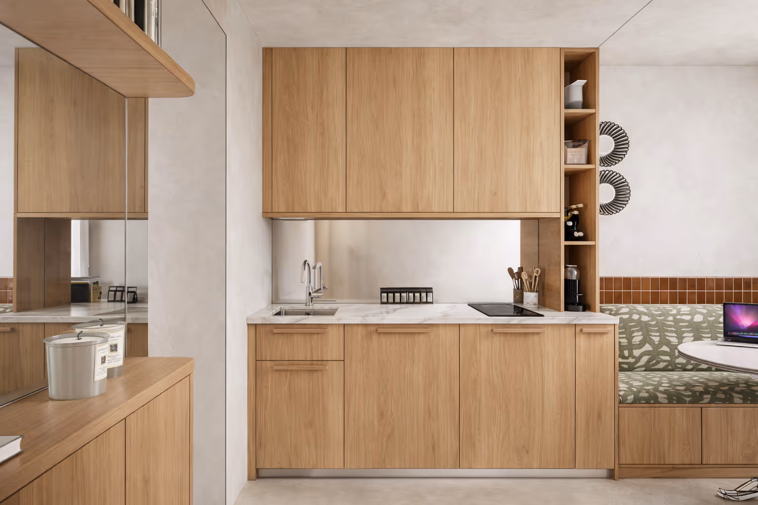 Cuisine moderne d'un studio parisien : placards en bois clair, plan de travail en marbre, évier et plaque de cuisson encastrés, coin repas avec banquette à motifs verts et table ronde. Réalisation : Aude Riou, architecte d'intérieur.